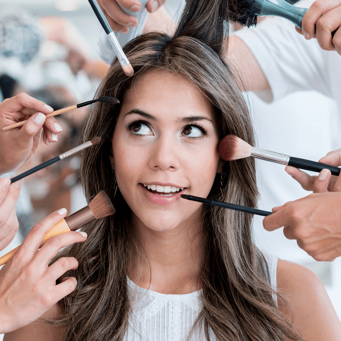Woman smiling while multiple hands apply makeup tools to her face and hair.